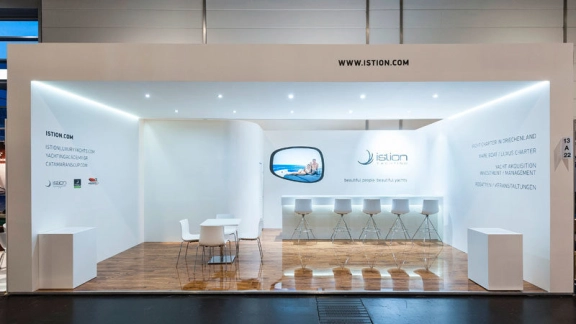 Heller, moderner Messestand mit weißem Tresen und Hockern, Tisch mit Stühlen, Holzfußboden, Bildschirm und Spotbeleuchtung.