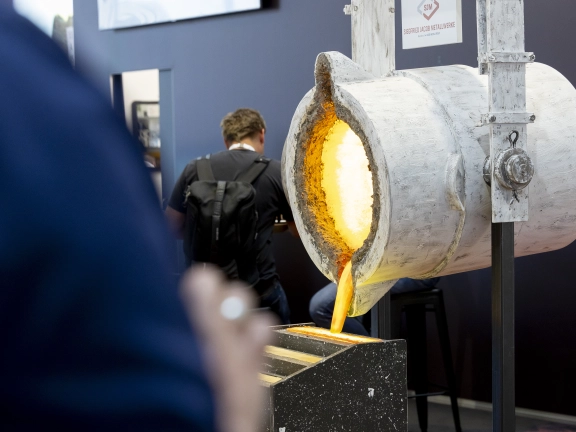 A crucible pours molten metal into a trough; a trade fair backdrop with people in the background.